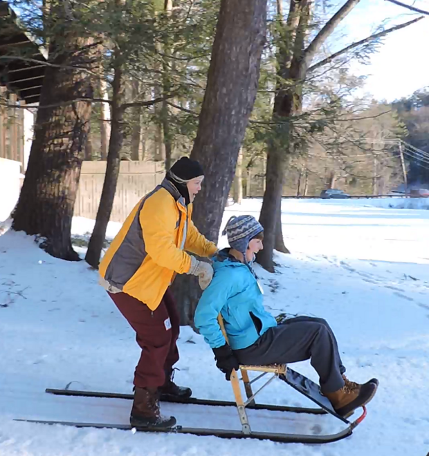 Two women are using a kicksled. The woman on the back is standing on the runners, while the woman in front sits in the chair. They are sledding down a hill and laughing.