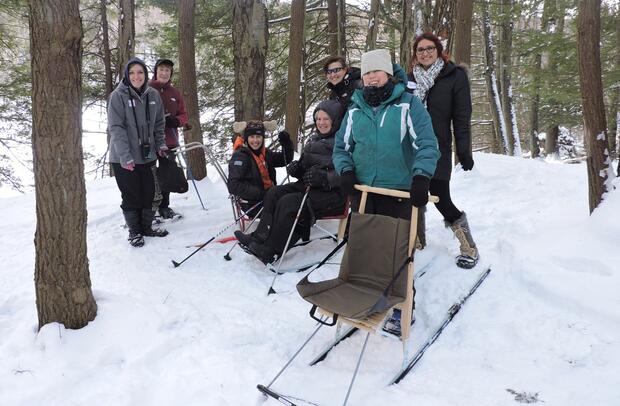 A group of people is standing in the snow in a forest. They are wearing warm winter clothing. Some of the people have grippers on their boots. One person is sitting in a sit ski with a push handle on the back of it. Another person is holding short ski poles and sitting in a kicksled. Everyone is smiling.