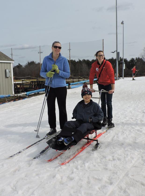 A standing skiier and a skiier in a sitski are next to each other in the snow. A woman is pushing the sit skier using handles on the back of the ski.