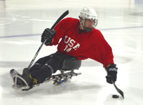 A sled hockey player wearing a mask and padding leans over to shoot a puck.