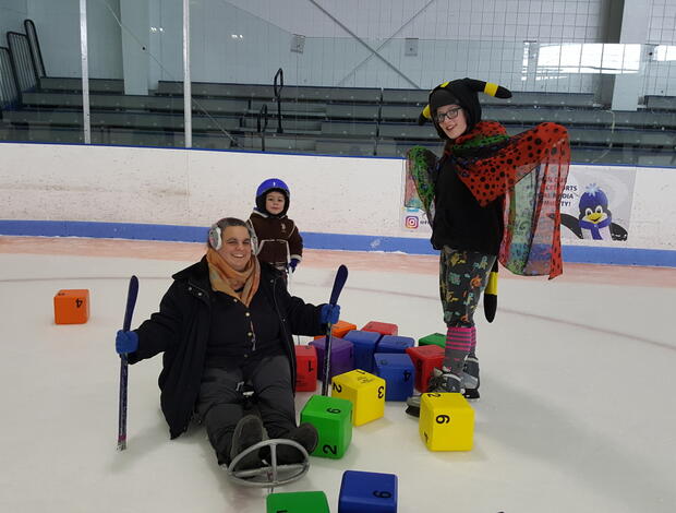 A woman in an ice sled, a girl on skates, and a young boy are standing in a pile of colorful blocks. They are smiling at the camera. The girl is spreading a colorful spotted cape with her arms, and wearing a hat with ears and a plush tail.