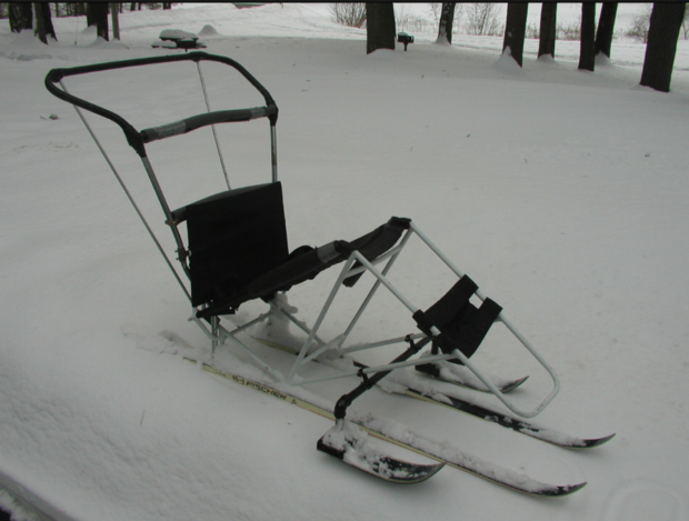 This sit ski has a frame that is bent up at the knees at a 110 degree angle. Two long skis are under this frame, and two smaller skis are on either side to act as outriggers. The sit ski has a padded seat and a strap for the skier's legs. A handle has been added to the back of the sit ski.