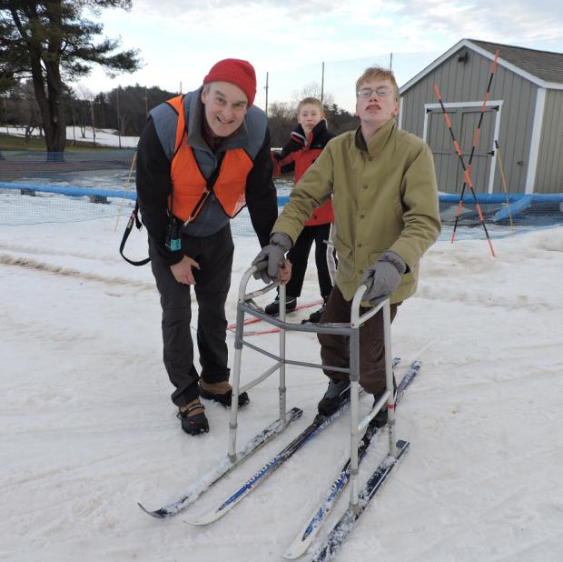 A skier is standing with bent knees, holding onto a walker that has skis attached to the bottom. A man witch cleats on his shows is standing next to the skier and smiling. Another skier is standing behind them.