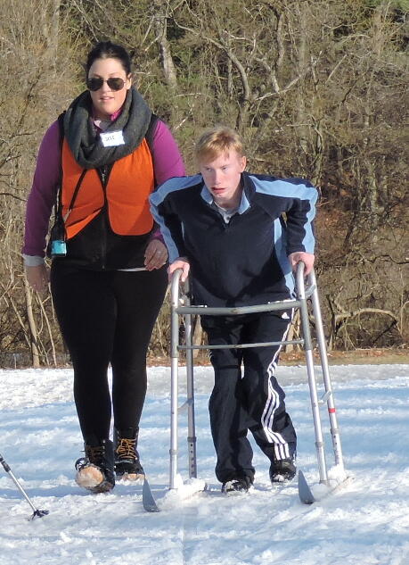A young man is pushing a walker in front of him in the snow. The walker has skis mounted on it. A young woman in an orange vest is walking beside him.