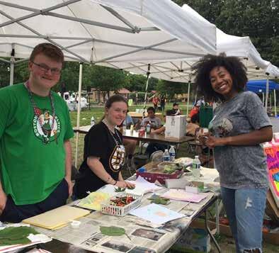 Three smiling people are standing in front of a table set up in a grassy field, covered with a canopy. The table is covered with art and activities. Several other tables and canopies are visible in the field behind them.