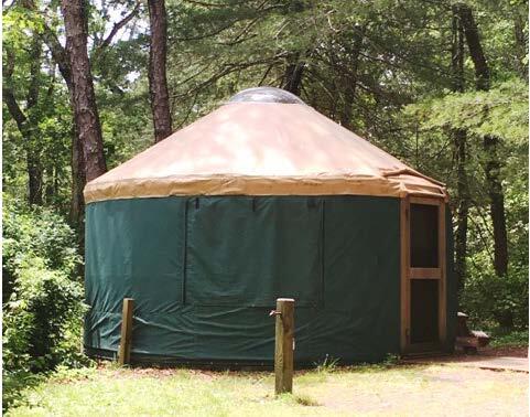 A round green yurt with a sloping tan roof sits in a grassy clearing. The yurt has a screen door. Woods are visible in the background.
