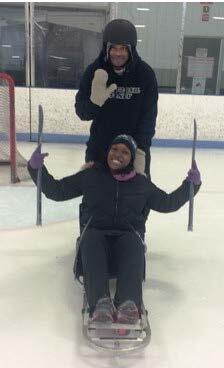 A man wearing a hockey helmet and mittens is standing behind a woman who is sitting in an ice sled. The man is waving. The woman is holding her hockey sticks in the air. They are both smiling.