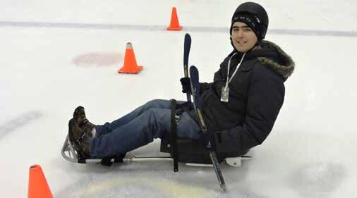 A skater wearing a warm winter coat is sitting in an ice sled. The skater is wearing a helmet, and holding two short hockey sticks. Cones are set up on the ice near the skater.