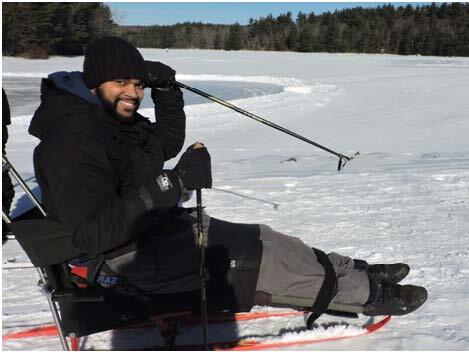 A man is sitting in a sit ski with his legs stretched out straight in front of him, and held in place with straps. The skier is holding two short ski poles, one of them raised in the air. A frozen lake is visible behind the skier.