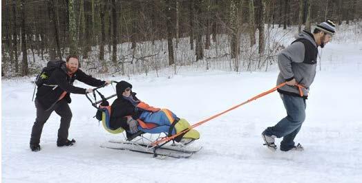 A woman is sitting in a ski with outriggers, wrapped in a sleeping bag. A man behind the skier is pushing her with handles on the back of the sit ski. Another man in front is pulling the skier with straps attached to the sit ski.