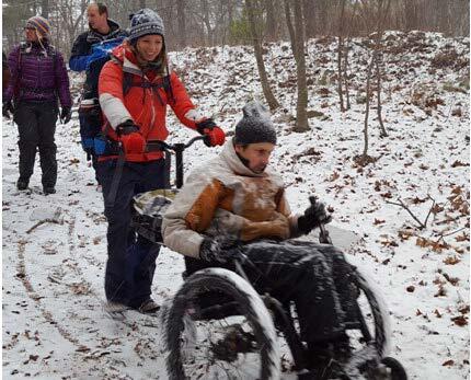 A hiker is using a wheelchair with levers and rugged tired to move through woods dusted lightly with snow. Another hiker is assisting with a push behind the chair. Several other hikers are visible behind them.