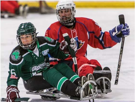 Two sled hockey players are next to each other on the ice. One of the players is wearing a green uniform and the other is wearing a red uniform. Both players are wearing helmets with face masks, shoulder pads, and large gloves. The players are looking off to the right.