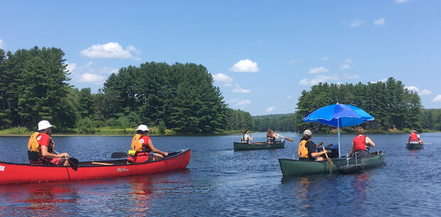 A group of canoes are moving past a wooded island. One of the canoes has pontoons and an umbrella on it. Another canoe has a seat with back and side support. The shore is lushly green and covered in trees.