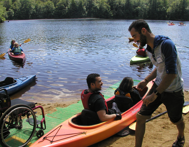 A tandem kayak sits on the beach, next to a run of beach mat. A wheelchair is sitting on the bach mat. Two people are sitting in the tandem kayak, facing the water. A man is walking towards the back of the kayak. Several other kayakers are paddling in the water.