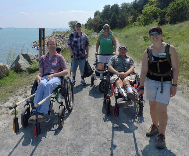 Five hikers are standing on a smooth dirt path. The ocean is on one side of them and a grassy, sloping hillis on the other. Two of the hikers are sitting in wheelchairs with rugged tires and rickshaw poles attached to the front. One hiker is using a cane, and another is using an all-terrain walker with three wheels. One of the hikers in a wheelchair is being pulled by a young woman with a volunteer name tag.