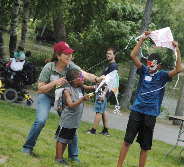 A woman and two children are standing on a grassy lawn, holding colorful kites with streamers. The children are wearing masks that have been colored with markers. One of the children is holding their kite above their head. Behind them is a path in front of a river. A person using a power wheelchair and a standing person are waking along the path.