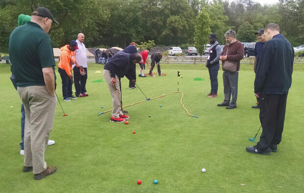 A group of about a dozen people is standing on a putting green. There is a rope on the green laid out in a V shape, with the hole at the tip of the V. Golfers are putting into the rope area using colorful golf clubs and golf balls.