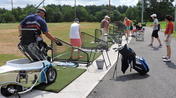 A group of people is standing on the driving range, driving golf balls. Several other people are standing behind them, watching and offering advice. one of the golfers is using a ParaGolfer to hold him upright while he swings. The reflection of a wheelchair, a service dog, and another ParaGolfe user are visible in the side of the ParaGolfer.
