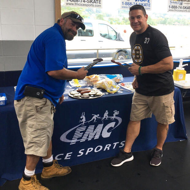 Two men are standing in front of a table with food on it. The man on the left is holding a hamburger in tongs, and the man on the right is holding a hot dog. The tablecloth reads FMC ICE SPORTS.