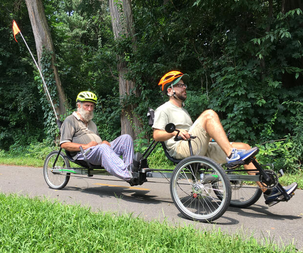 Two men are riding a tandem recumbent tricycle. The man in front is steering. The man in back has a long white beard, and is sitting with his hands in his lap.