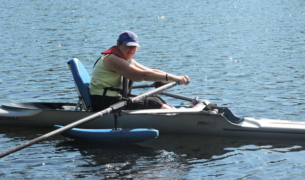 A woman is rowing in a shell that has pontoons on either side. She is sitting in an upright seat with a high back.