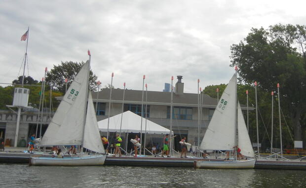Two sailboats are tied up on a long dock. Several people are walking along the dock. There is a stone building in the background.