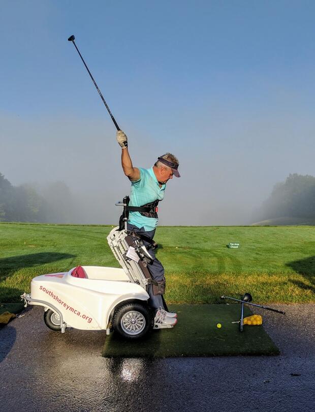 A man is using a Paragolfer to raise himself to a standing position. He is swinging his club high in the air to swing at a golf ball on the ground in front of him.