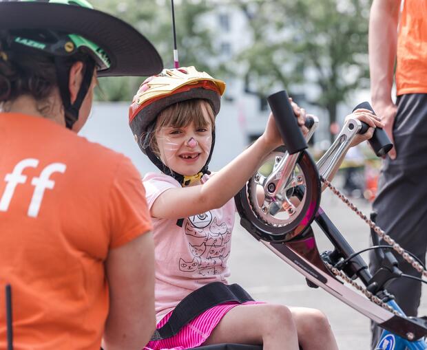A young girl is seated in a trike holding the handlebars with a seat buckle around her waist. There are 2 staff members to her left and right giving her instructions.