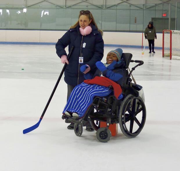 A participant, bundled up warm winter clothes, is sitting in her wheelchair on the ice and laughing. A woman to her right is holding hockey stick and wearing ice grippers on her shoes.