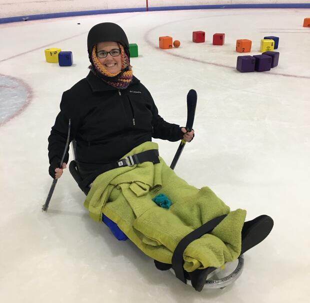 A participant is sitting in a ice sled with a blanket wrapped around her legs. She has straps holding her legs onto the sled's footrest. She is holding short hockey sticks and has a big smile on her face. The colorful blocks are visible behind her.