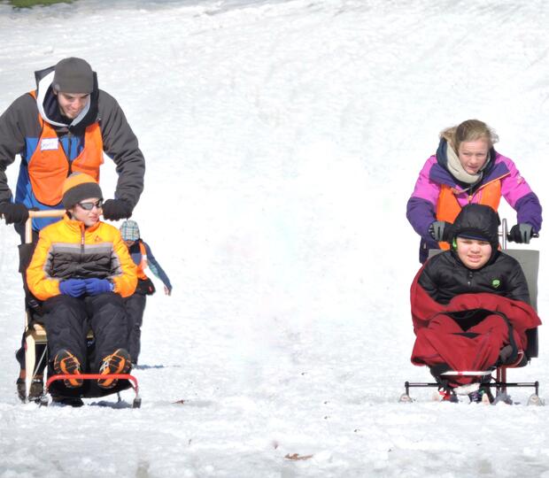 A kicksled user and a sit-skier are being pushed down a hill by volunteers.