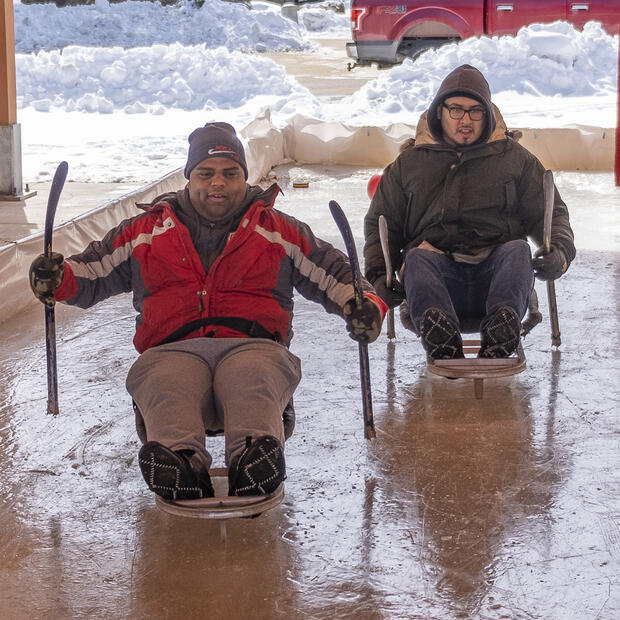 Two people are sitting in ice sleds the outdoor skating rink. They are each holding short hockey sticks and gripping the ice to glide around the rink.