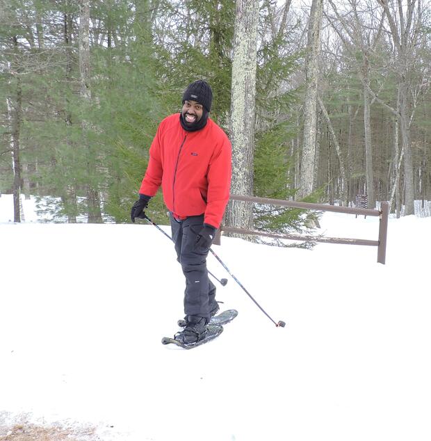 A man dressed in warm winter clothes has snowshoes on and is holding ski poles for balance. He is flashing a big smile as he snowshoes up the hill on a trail.