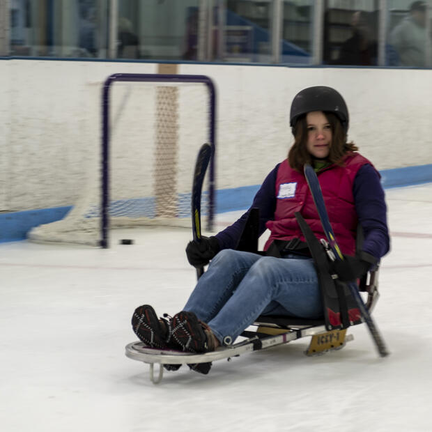 A woman is sitting in an ice sled and holding short hockey sticks to propel herself on the ice.