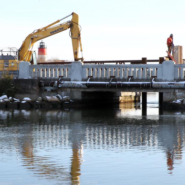 A person an a backhoe on a small bridge with a lighthouse in the background.