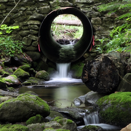 Water flowing out of a metal pipe culvert surrounded by rock.