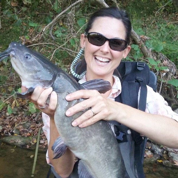 Jenny holding a catfish while standing in a stream and smiling at the camera.