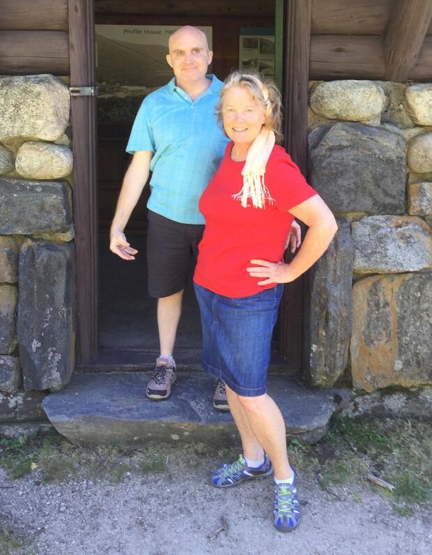 Glenn and Cheryl standing outdoors together in front of the door to a log cabin