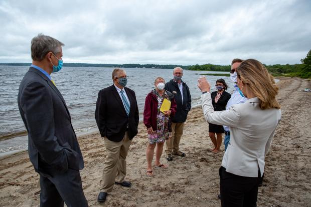 A group of people, including Governor Baker and partners, speaking in a circle on a beach.
