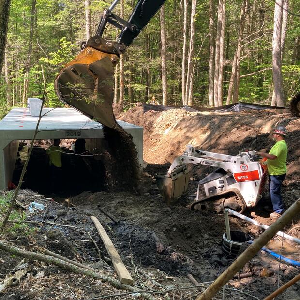 A person operating heavy machinery and digging near a new and exposed cement culvert.