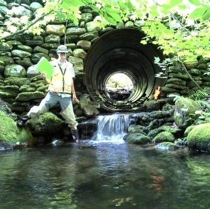 A person standing in a stream holding a clipboard in front of a culvert with water flowing out of it.