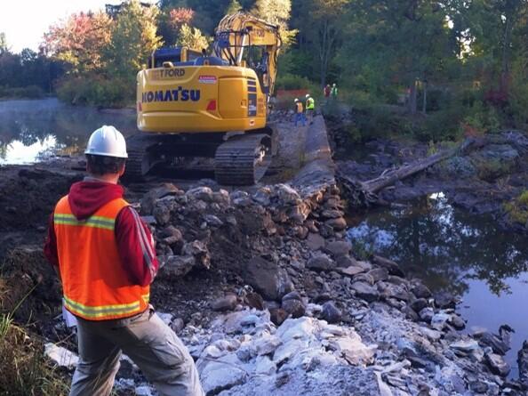 A person in a hard hat and high visibility vest standing near construction equipment on a dam with water on either side.