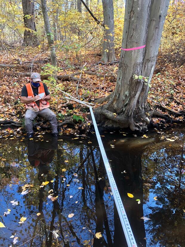 A person writing on a clipboard while sitting on the bank of a stream.