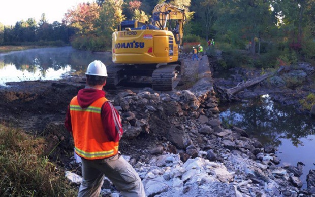 A person standing on the shore of a river and looking at an excavator on a partially-removed dam.