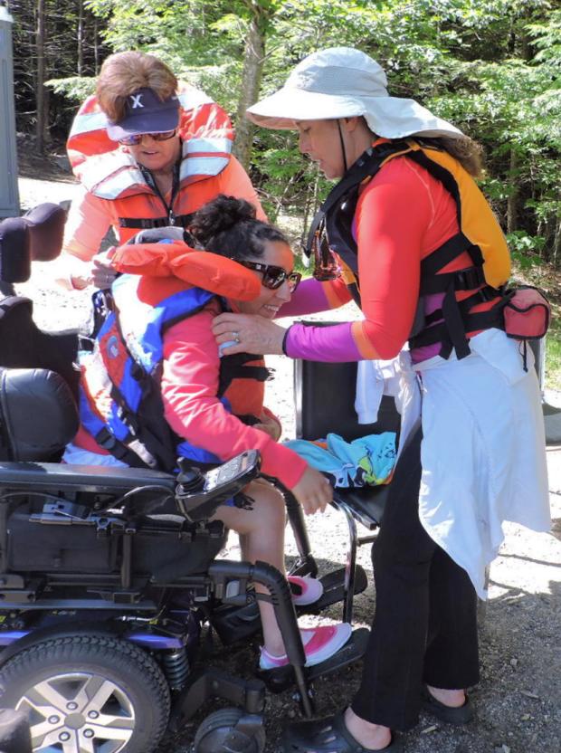 A paddler using a power wheelchair is fitted with a PFD by a staff person and a companion.