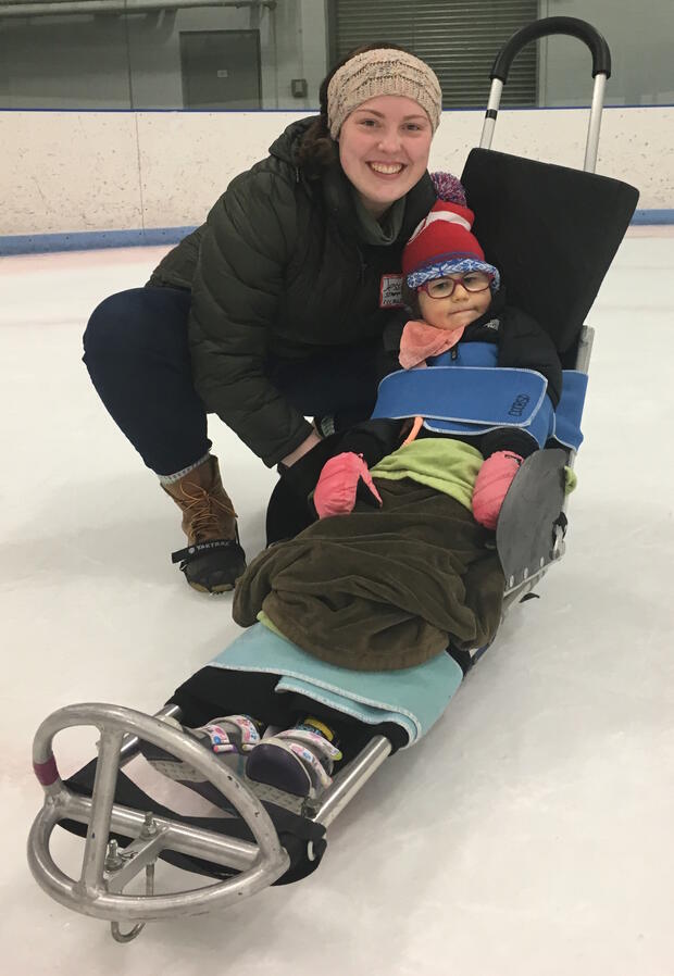 An adult is crouching down next to a child in an ice sled. The child is wrapped in blankets and strapped into the sled. Foam wedges are behind the child's back and head.