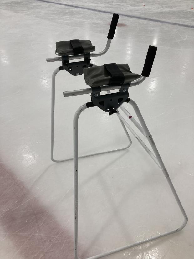 A metal walker without wheels. The sides of the walker are shaped like tall trapezoids, with sides of equal height and a bottom that is wider than the top. On the top are two handles shalede like Ls lying on their backs with the short end sticking up in the air. There is padding and a strap in the middle of the long arm of the L.