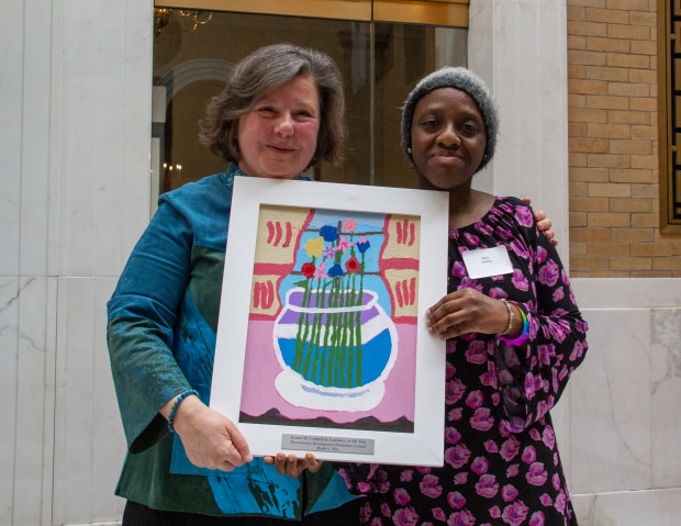 Senator Comerford, a caucasian woman with light brown and gray hair wearing a blue and green shirt stands holding a white framed painting of a vase of flowers with Betty Antoine, a Black woman wearing a black and pink floral patterned shirt and a gray beanie