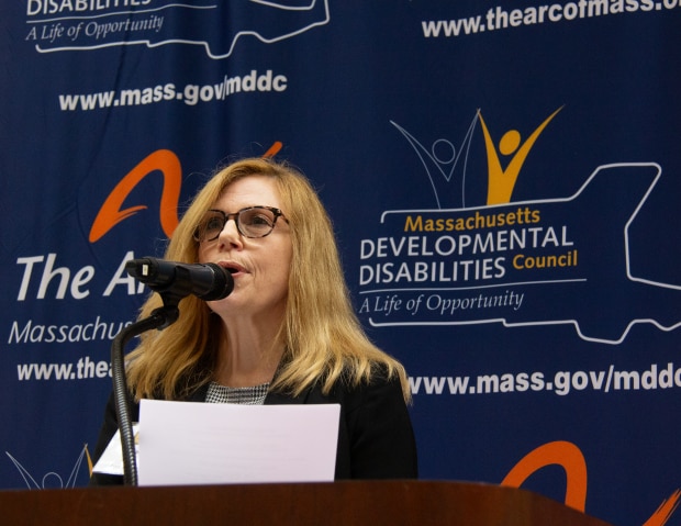 Heidi Robbins, a caucasian woman with blonde hair wearing a black blazer and glasses stands speaking at a podium.