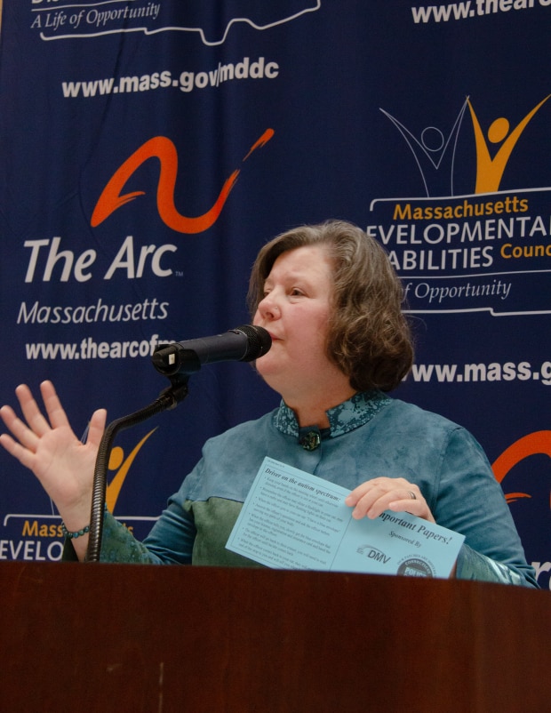 Senator Comerford, a caucasian woman with light brown and gray hair wearing a blue and green shirt stands at a podium speaking while holding a blue envelope.